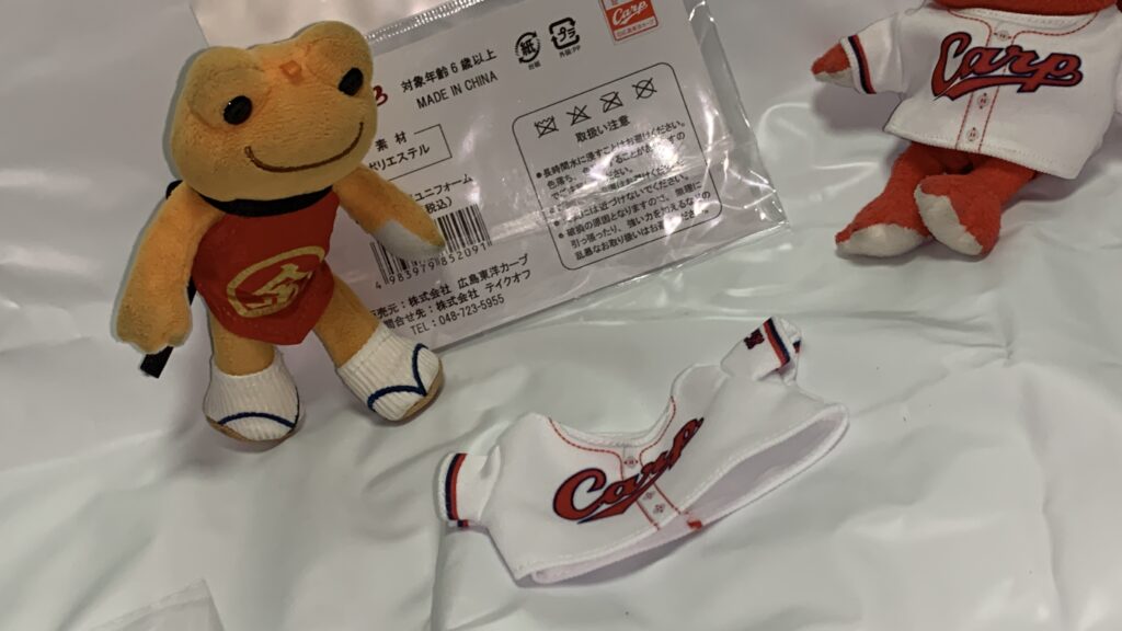 開封した白いカープのぬいぐるみ用ユニフォームと、着用前のオレンジ色のマスコットピクルス